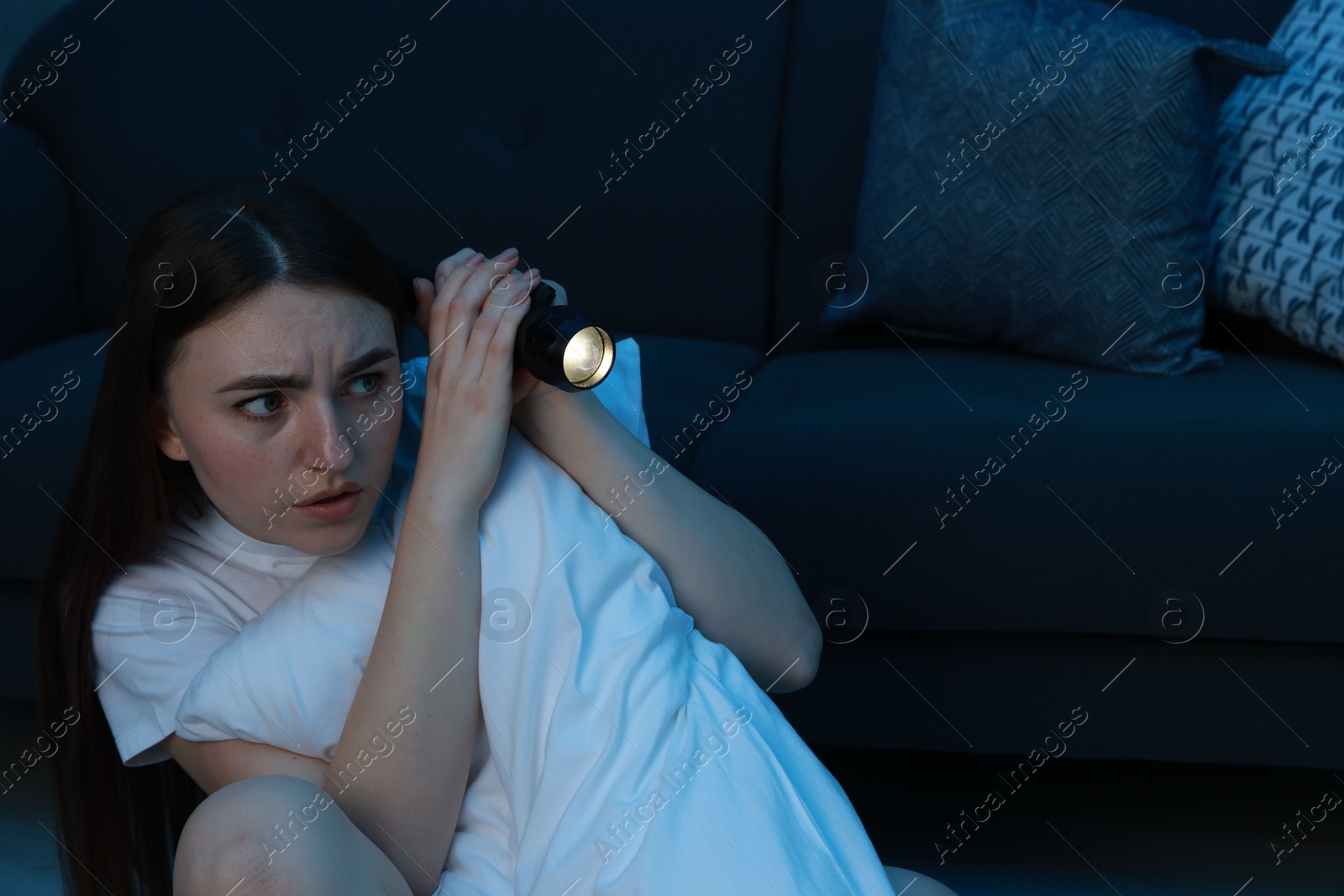 Photo of Fear of darkness. Scared young woman with flashlight hugging pillow near couch indoors at night, space for text