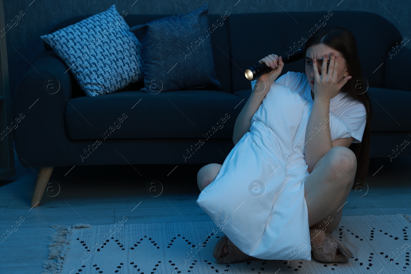 Photo of Fear of darkness. Scared young woman with flashlight hugging pillow and hiding her face near couch indoors at night, space for text