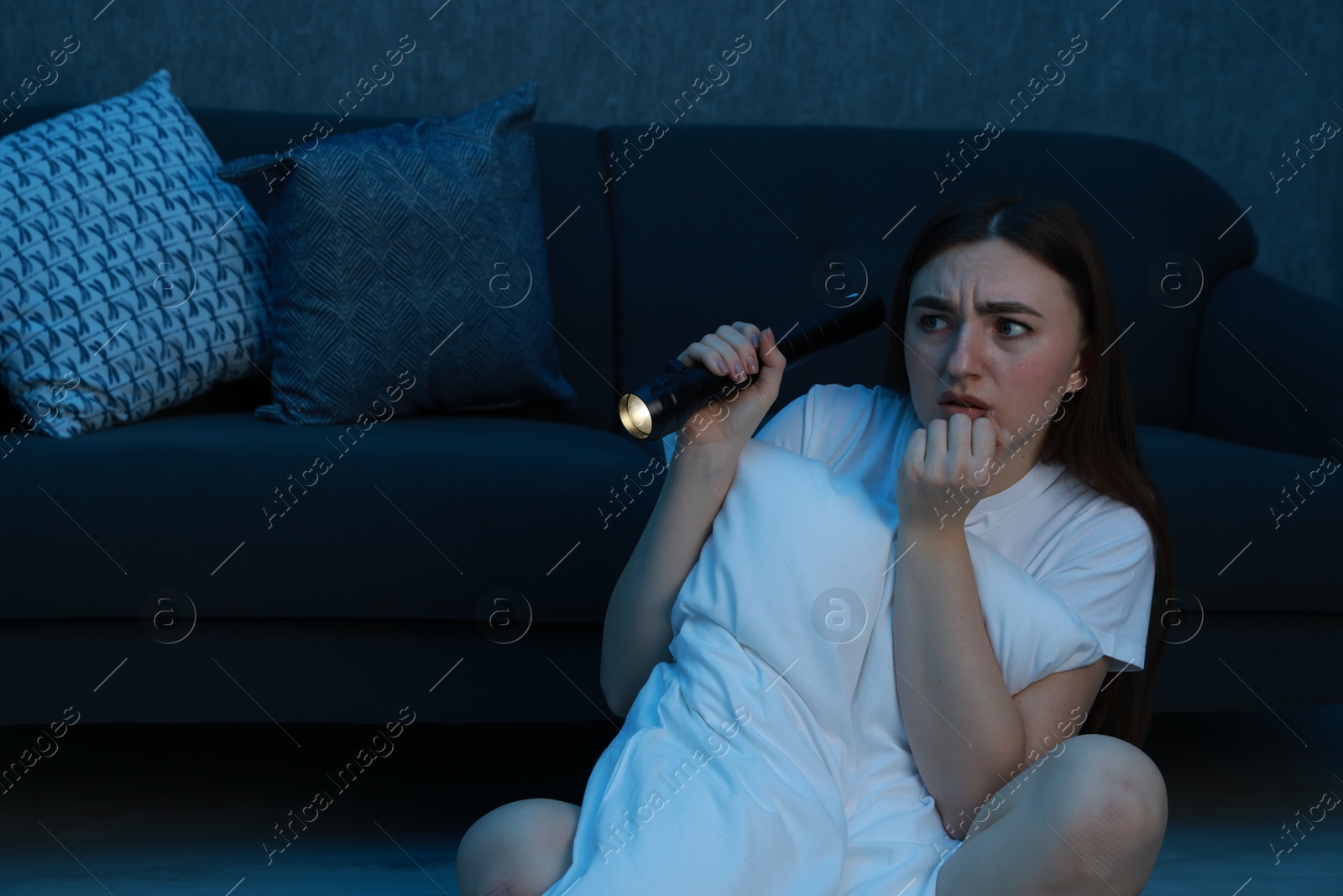 Photo of Fear of darkness. Scared young woman with flashlight hugging pillow near couch indoors at night, space for text