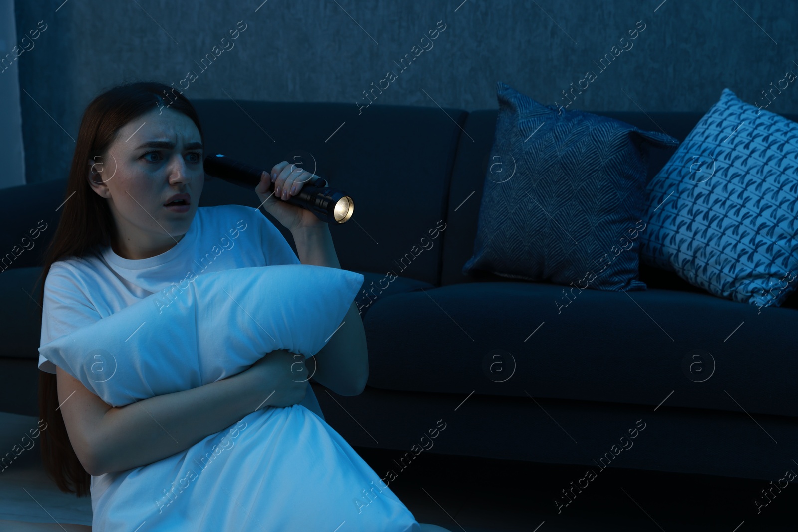 Photo of Fear of darkness. Scared young woman with flashlight hugging pillow near couch indoors at night, space for text