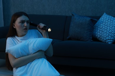 Photo of Fear of darkness. Scared young woman with flashlight hugging pillow near couch indoors at night, space for text