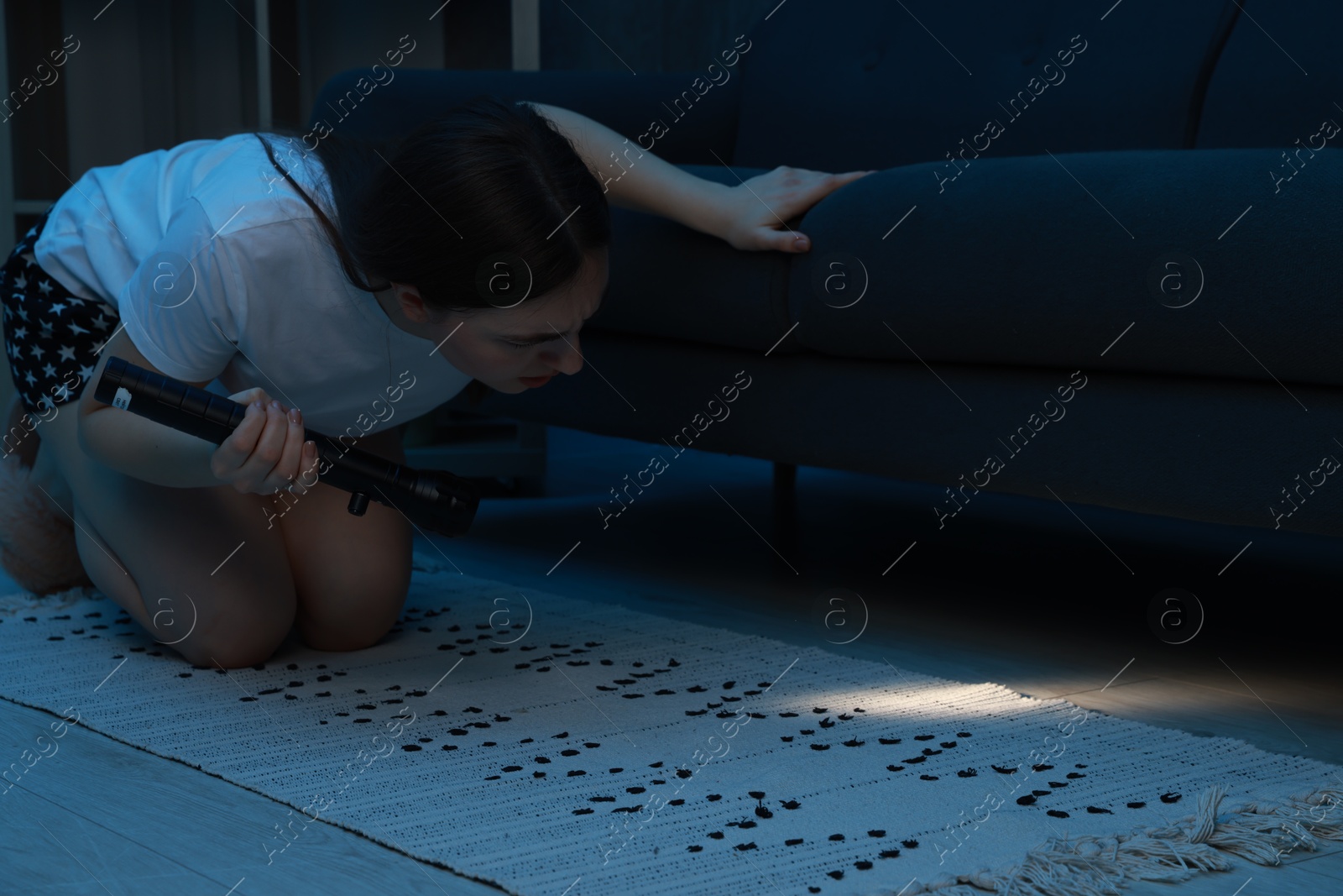 Photo of Fear of darkness. Scared young woman looking under couch with flashlight indoors at night, space for text