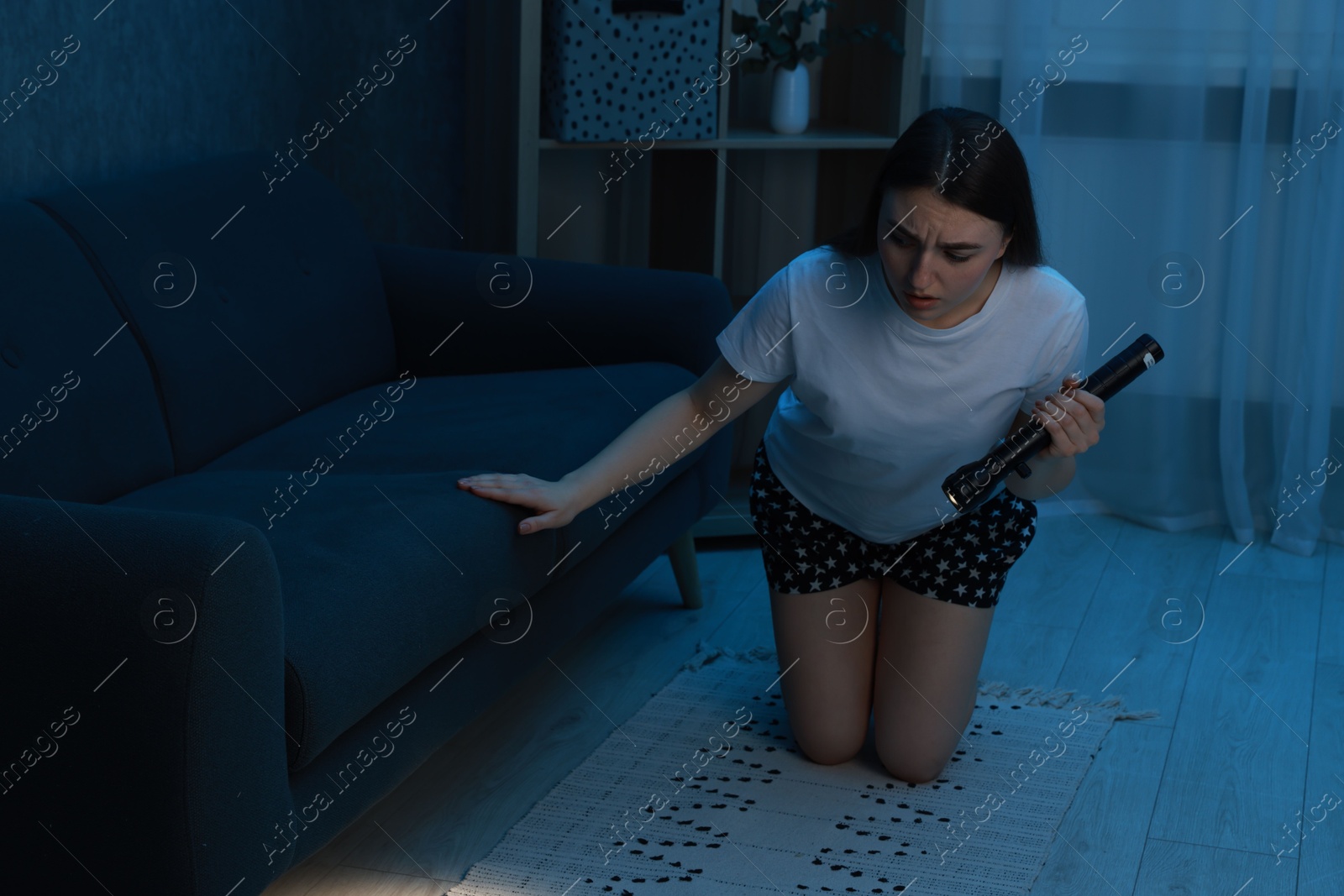 Photo of Fear of darkness. Scared young woman looking under couch with flashlight indoors at night