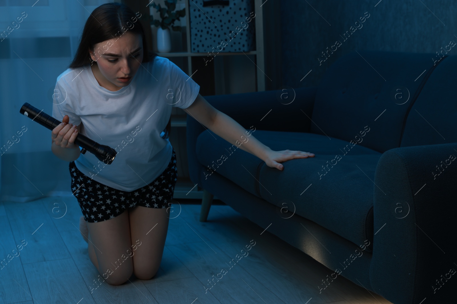 Photo of Fear of darkness. Scared young woman looking under couch with flashlight indoors at night