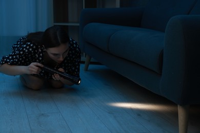 Photo of Fear of darkness. Scared young woman looking under couch with flashlight indoors at night, space for text