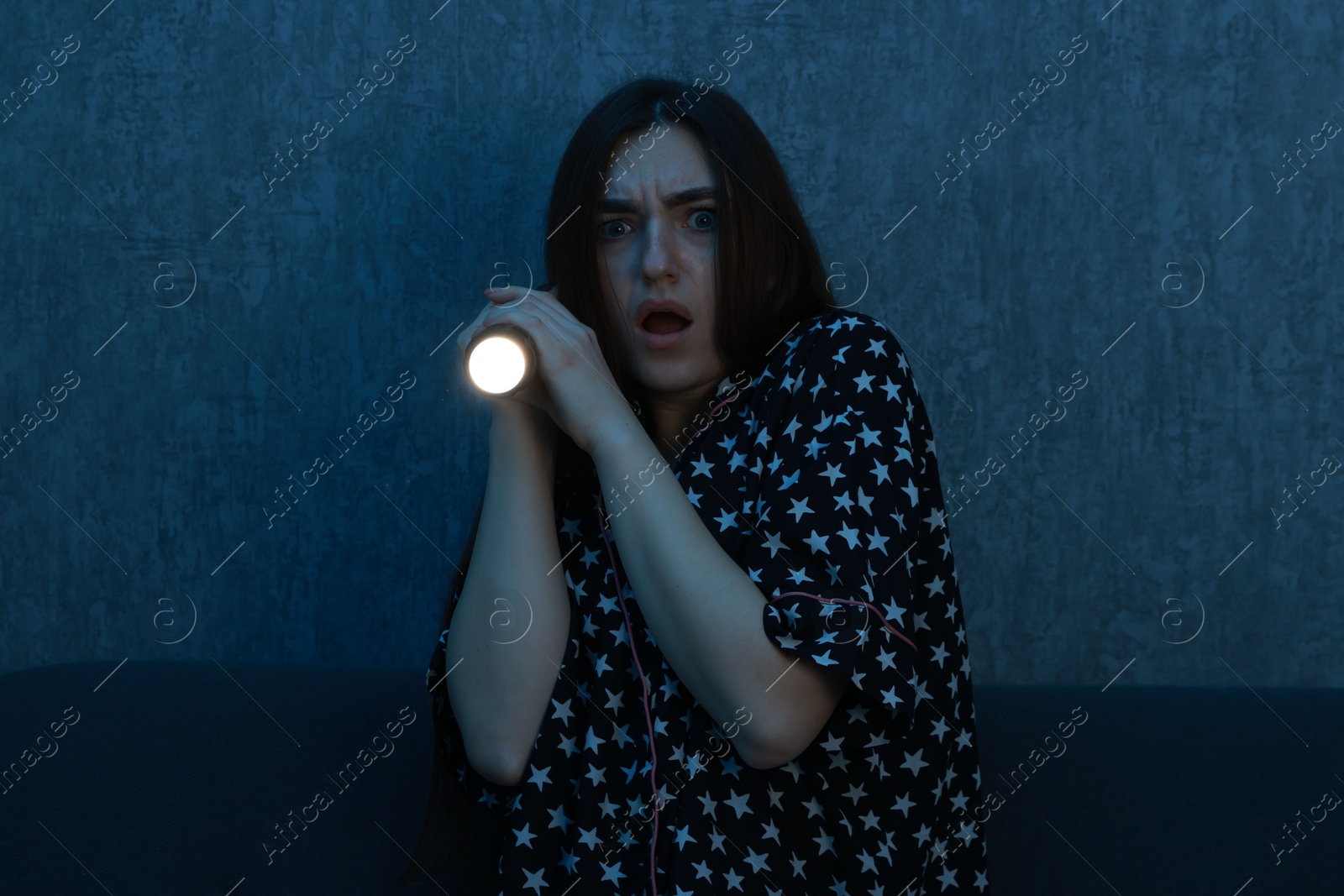 Photo of Fear of darkness. Scared young woman with flashlight on couch indoors at night