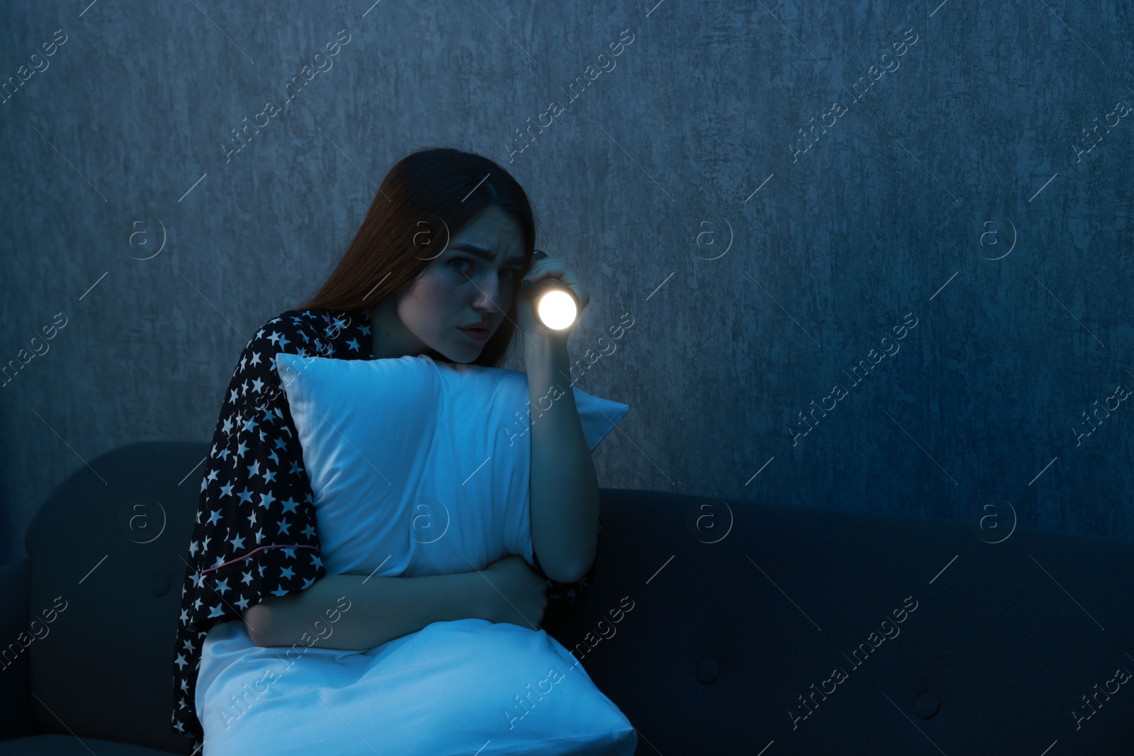 Photo of Fear of darkness. Scared young woman with flashlight hugging pillow on couch indoors at night, space for text