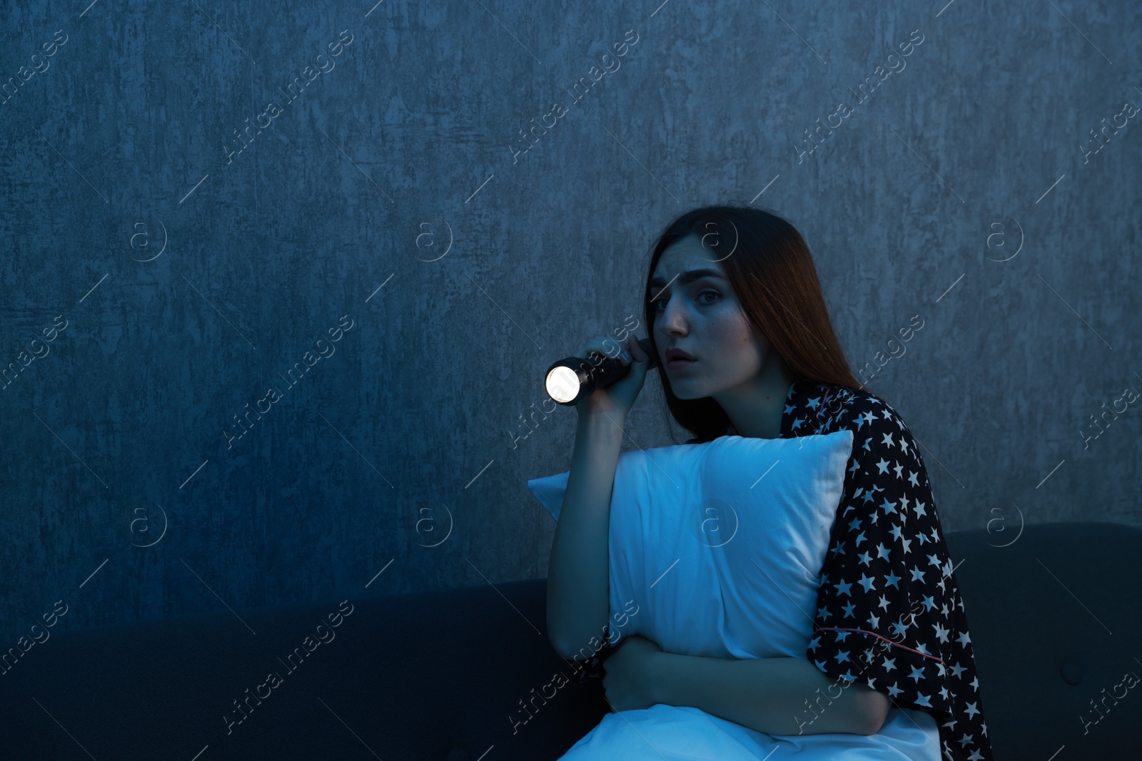 Photo of Fear of darkness. Scared young woman with flashlight hugging pillow on couch indoors at night, space for text