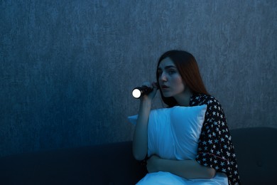 Photo of Fear of darkness. Scared young woman with flashlight hugging pillow on couch indoors at night, space for text