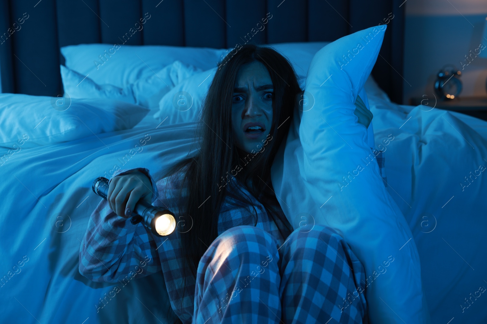 Photo of Fear of darkness. Scared young woman with flashlight hugging pillow in bedroom at night