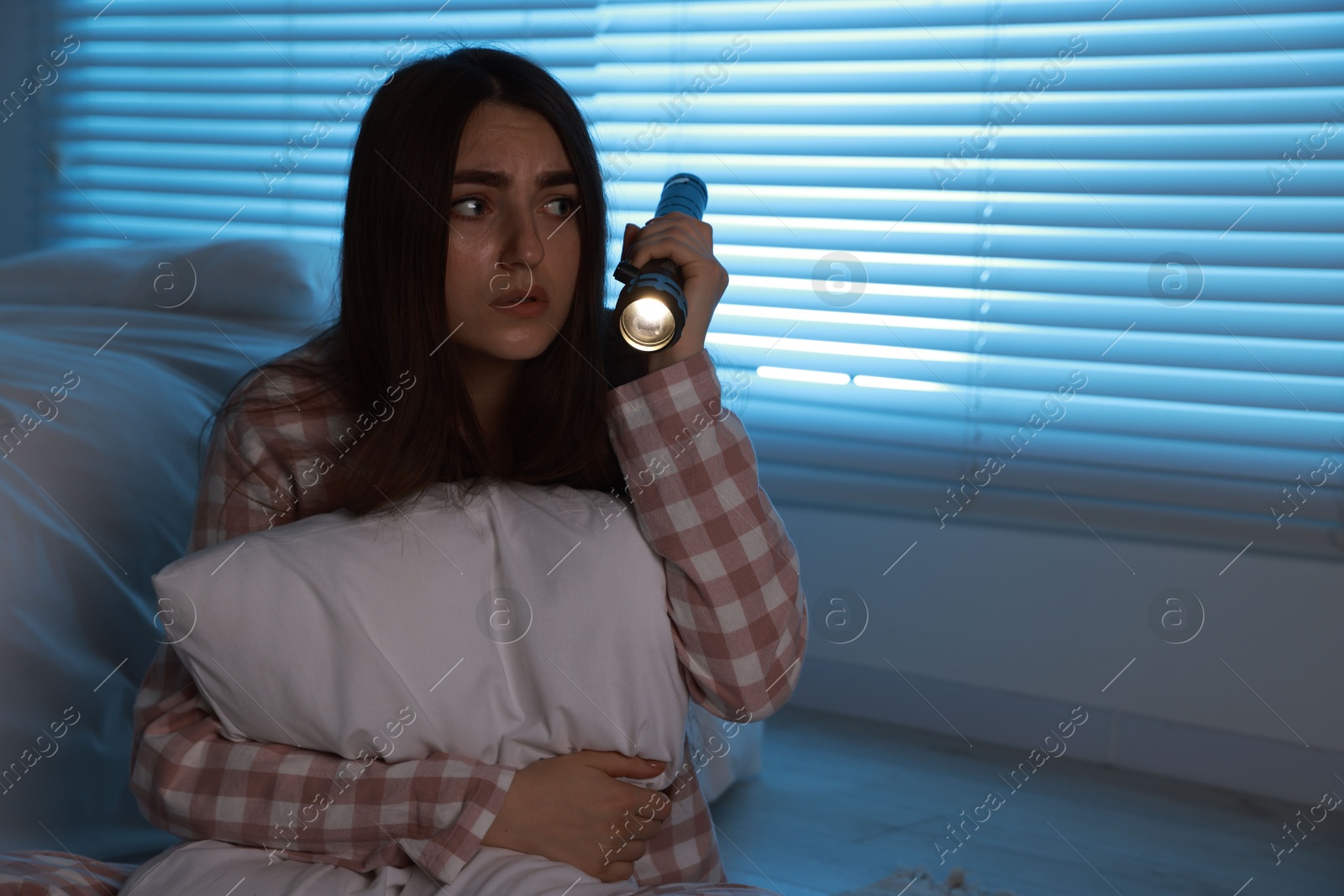 Photo of Fear of darkness. Scared young woman with flashlight hugging pillow in bedroom at night, space for text