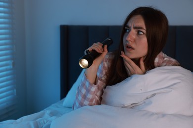 Photo of Fear of darkness. Scared young woman with flashlight lying on bed in room at night, space for text