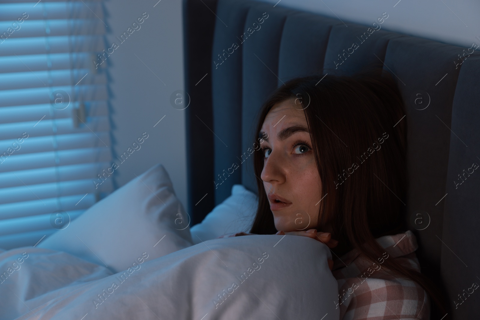 Photo of Fear of darkness. Scared young woman lying on bed in room at night, space for text