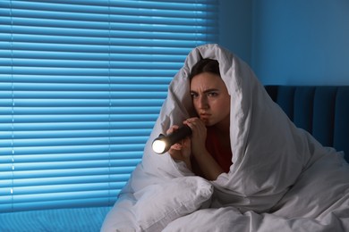 Photo of Fear of darkness. Scared young with blanket and flashlight on bed indoors at night