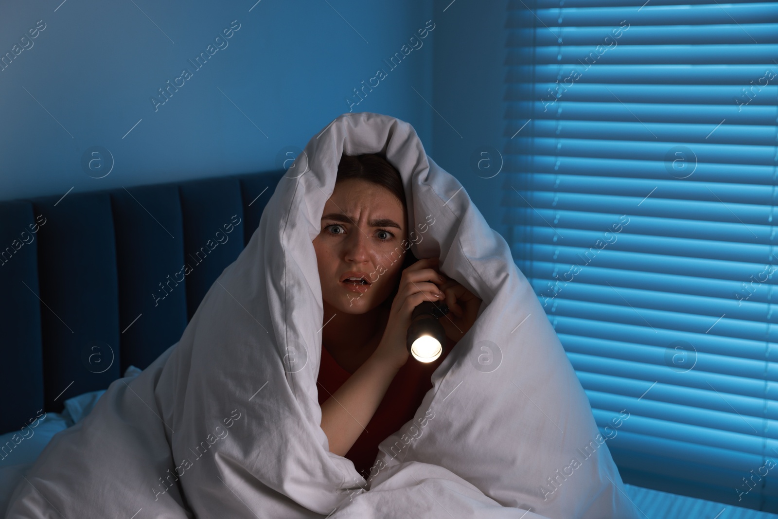 Photo of Fear of darkness. Scared young with blanket and flashlight on bed indoors at night