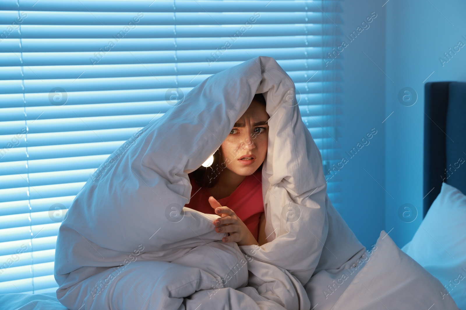 Photo of Fear of darkness. Scared young with blanket and flashlight on bed indoors at night