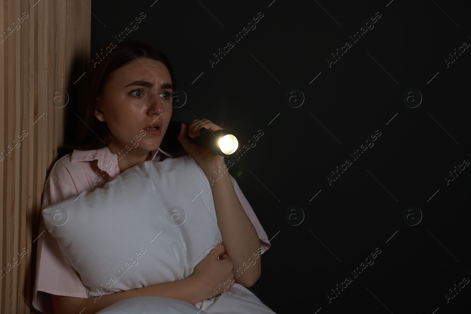 Photo of Fear of darkness. Scared young woman with flashlight hugging pillow indoors at night, space for text