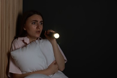 Photo of Fear of darkness. Scared young woman with flashlight hugging pillow indoors at night, space for text