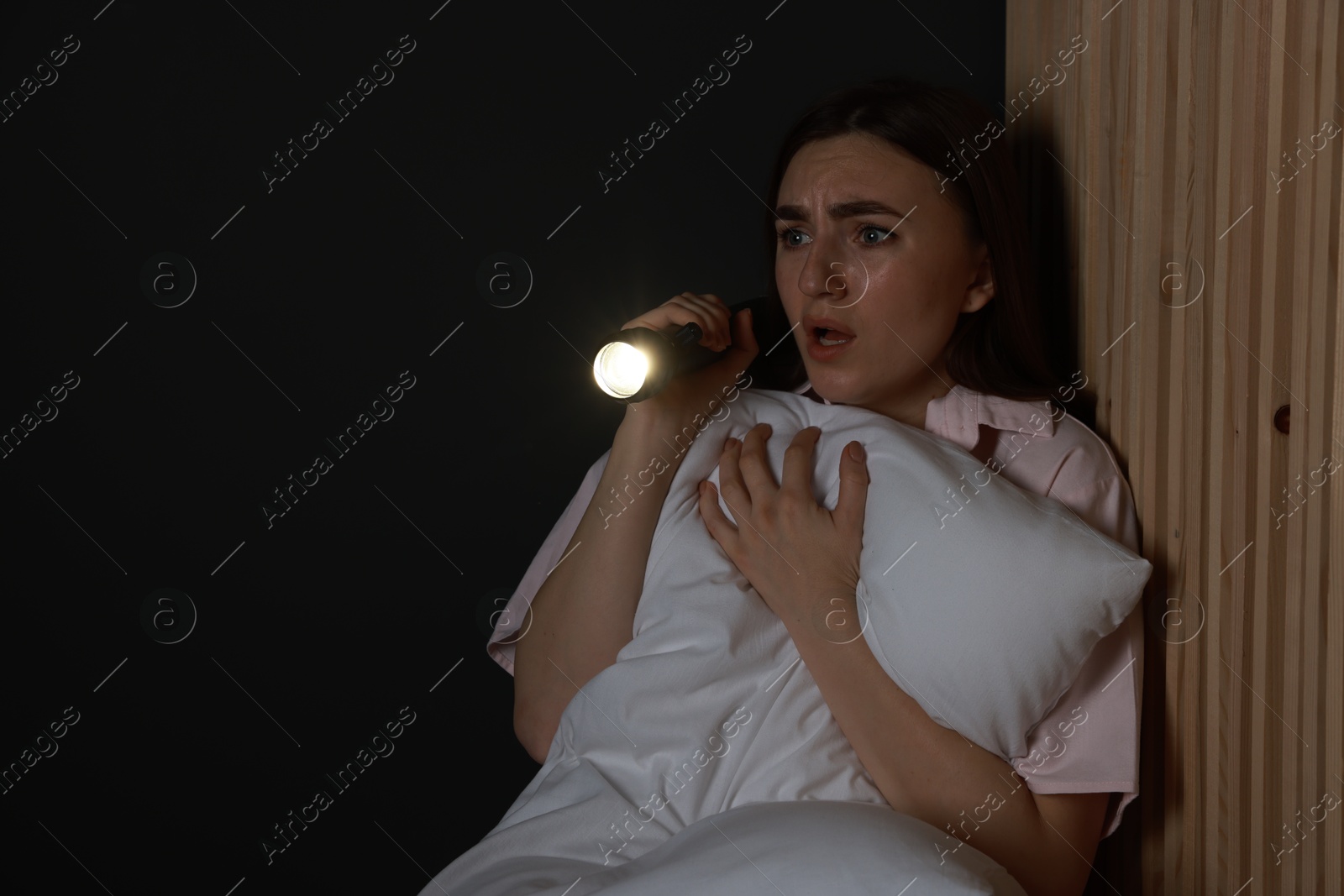 Photo of Fear of darkness. Scared young woman with flashlight hugging pillow indoors at night, space for text