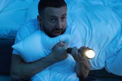 Fear of darkness. Scared man with flashlight hugging pillow in bedroom at night