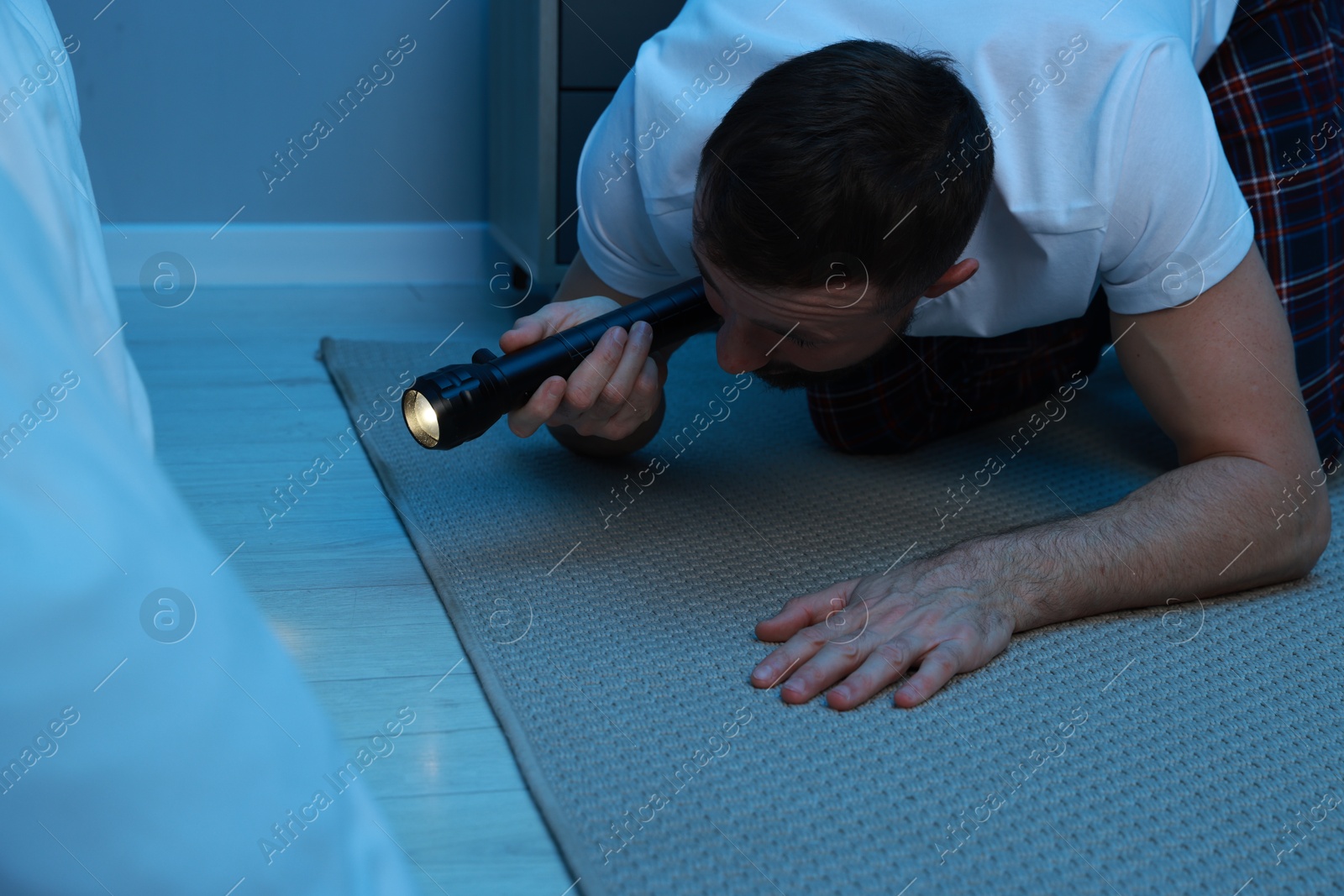 Photo of Fear of darkness. Scared man looking under bed with flashlight indoors at night