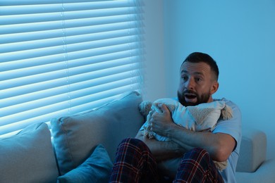 Fear of darkness. Scared man hugging pillow on couch in dark room, space for text