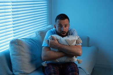 Fear of darkness. Scared man hugging pillow on couch in dark room