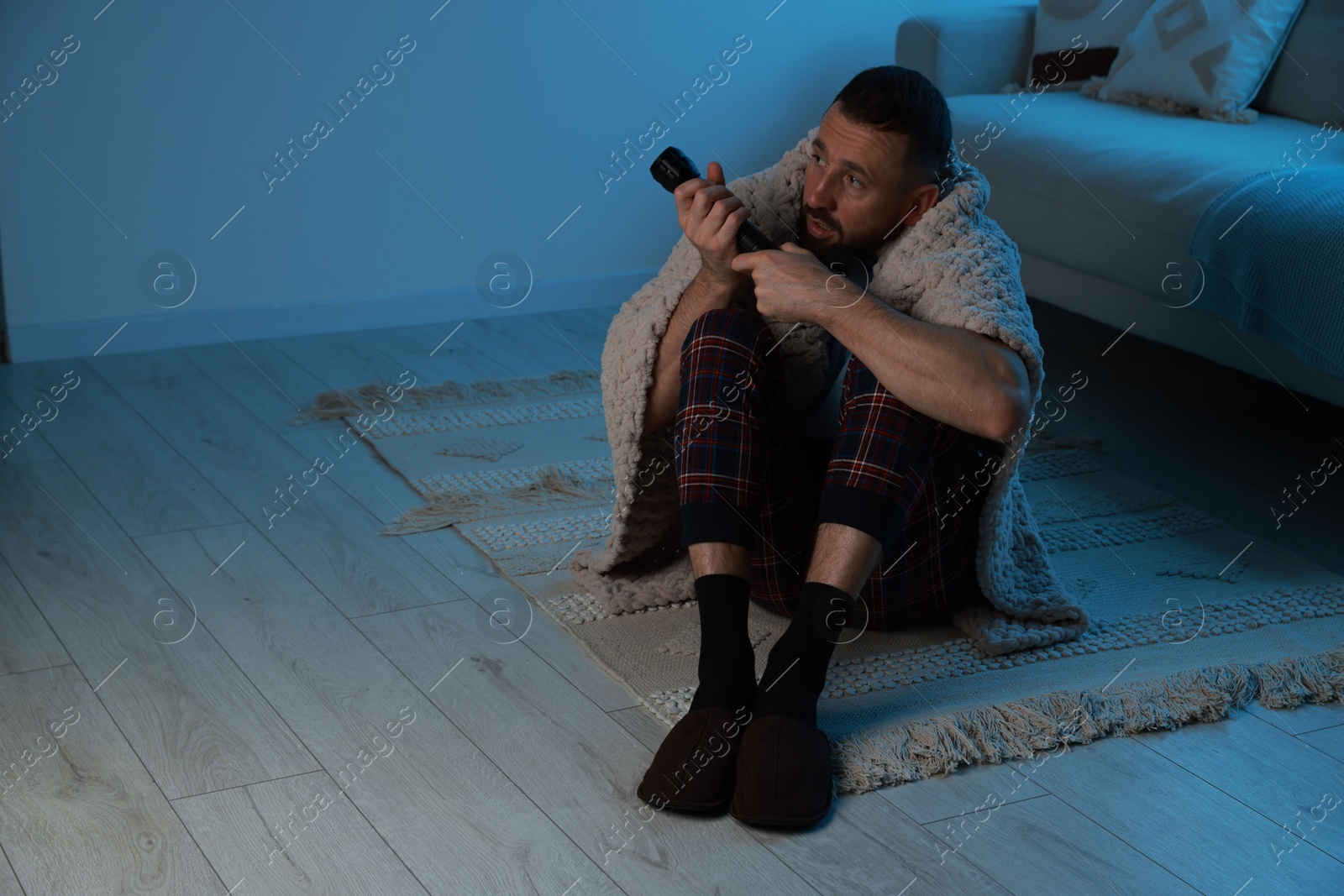 Photo of Fear of darkness. Scared man with blanket and flashlight indoors at night, space for text