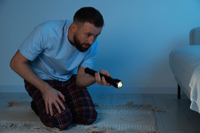 Photo of Fear of darkness. Scared man looking under bed with flashlight indoors at night