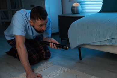 Photo of Fear of darkness. Scared man looking under bed with flashlight indoors at night