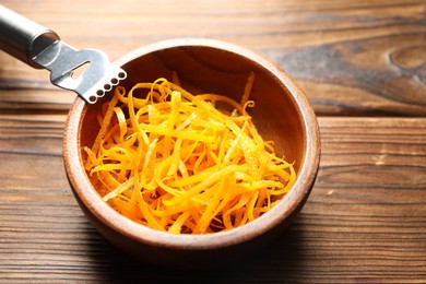 Photo of Fresh orange zest in bowl and zester tool on wooden table, closeup