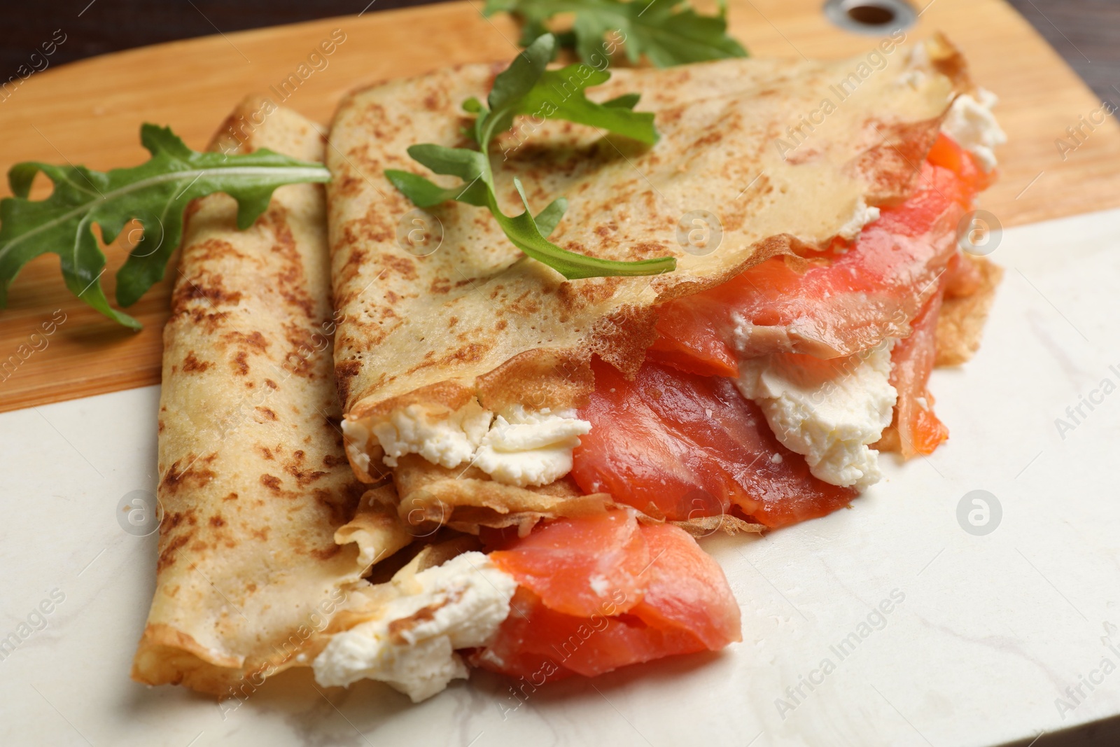 Photo of Delicious crepes with salmon, cream cheese and arugula on wooden board, closeup