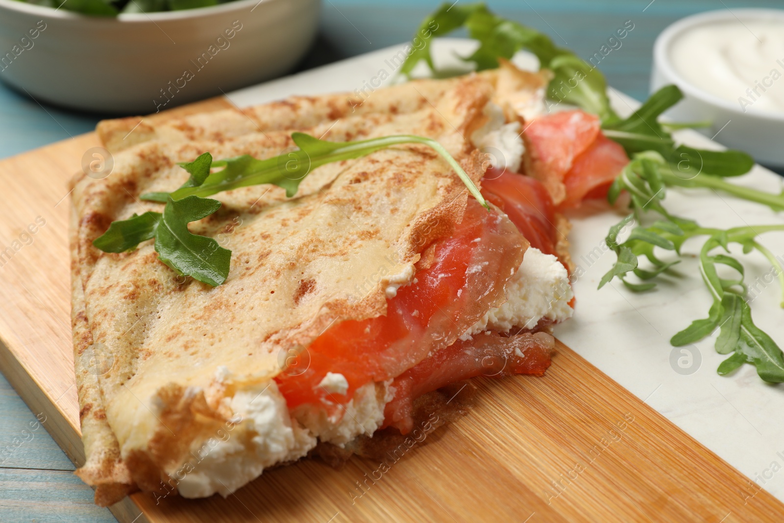 Photo of Delicious crepes with salmon, cream cheese and arugula on blue wooden table, closeup