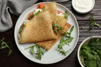 Photo of Delicious crepes with salmon, cream cheese and arugula on wooden table, flat lay