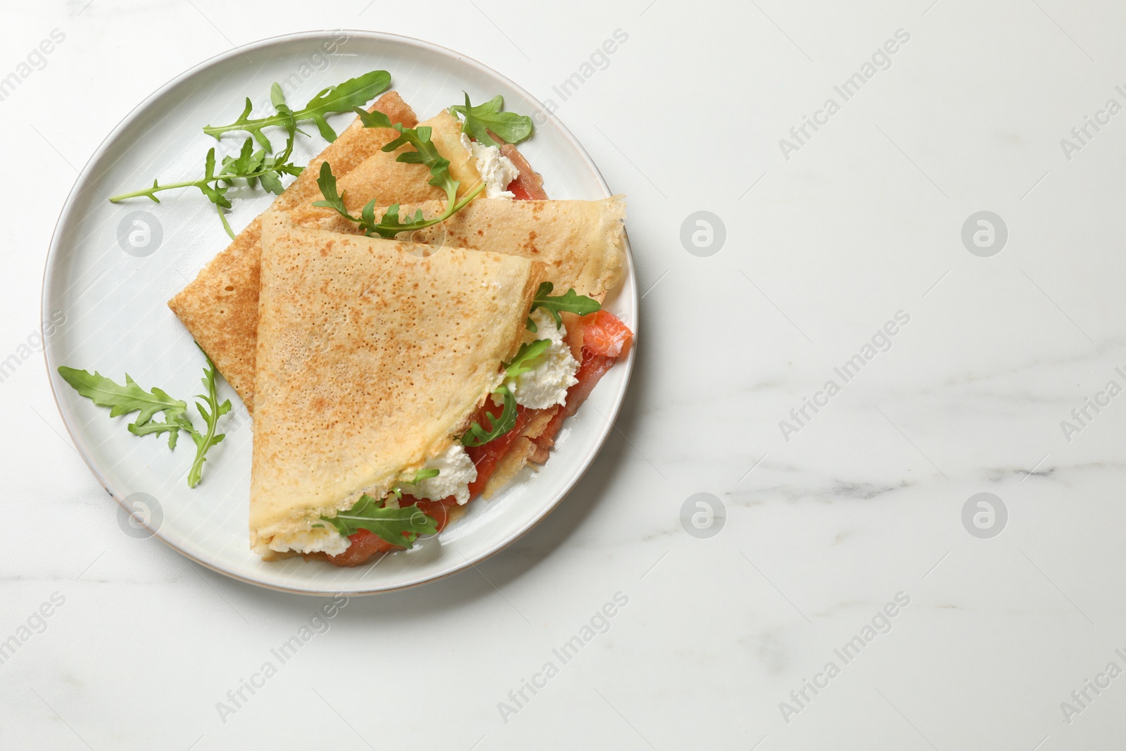 Photo of Delicious crepes with salmon, cream cheese and arugula on white marble table, top view. Space for text