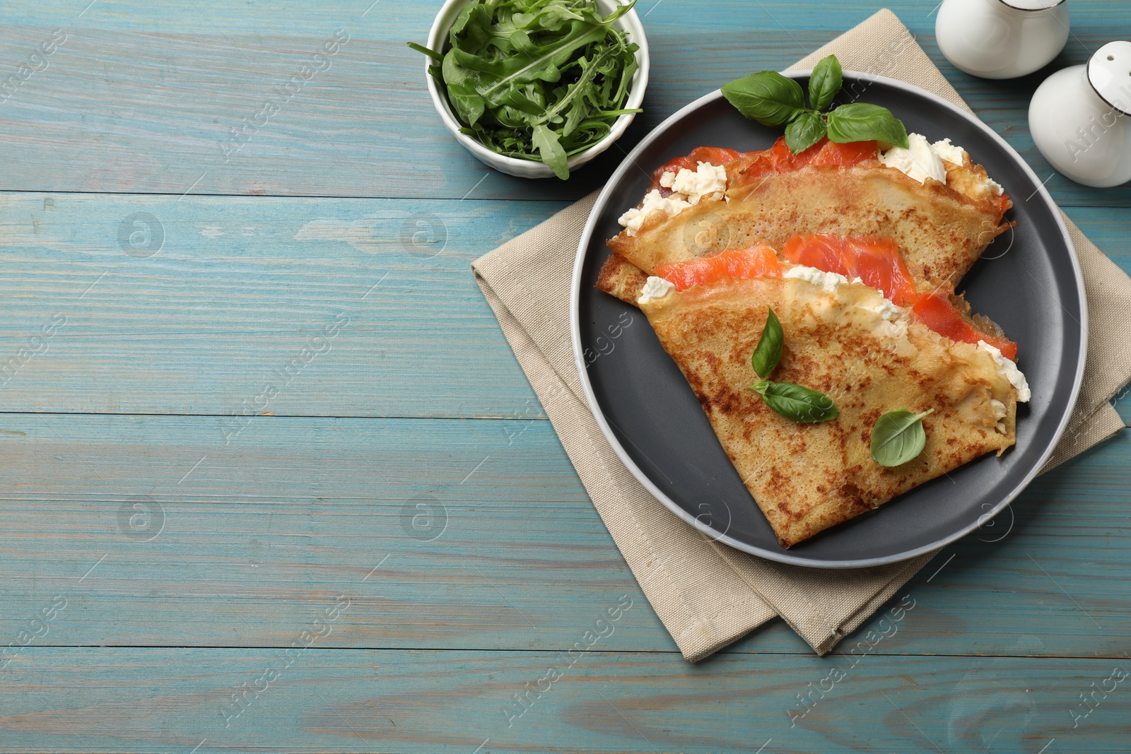 Photo of Delicious crepes with salmon, cream cheese, basil and arugula on light blue wooden table, flat lay. Space for text