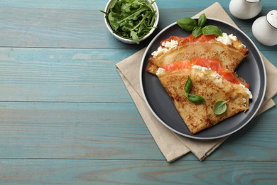 Photo of Delicious crepes with salmon, cream cheese, basil and arugula on light blue wooden table, flat lay. Space for text