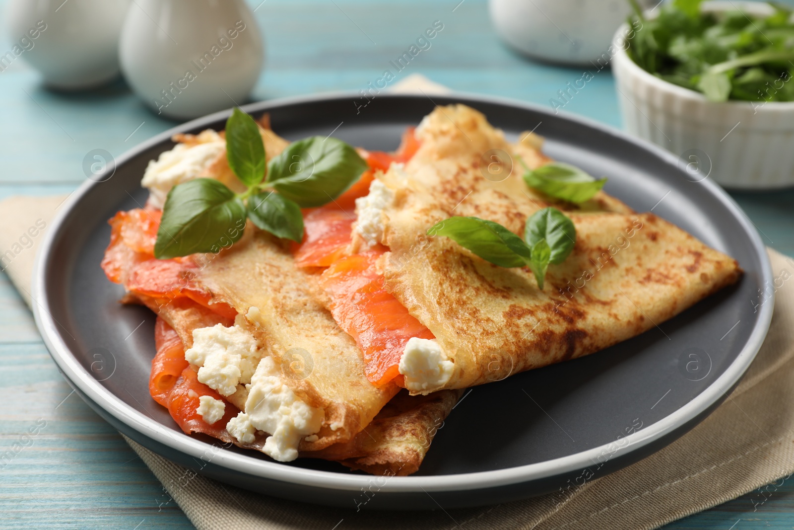 Photo of Delicious crepes with salmon, cream cheese and basil on light blue wooden table, closeup