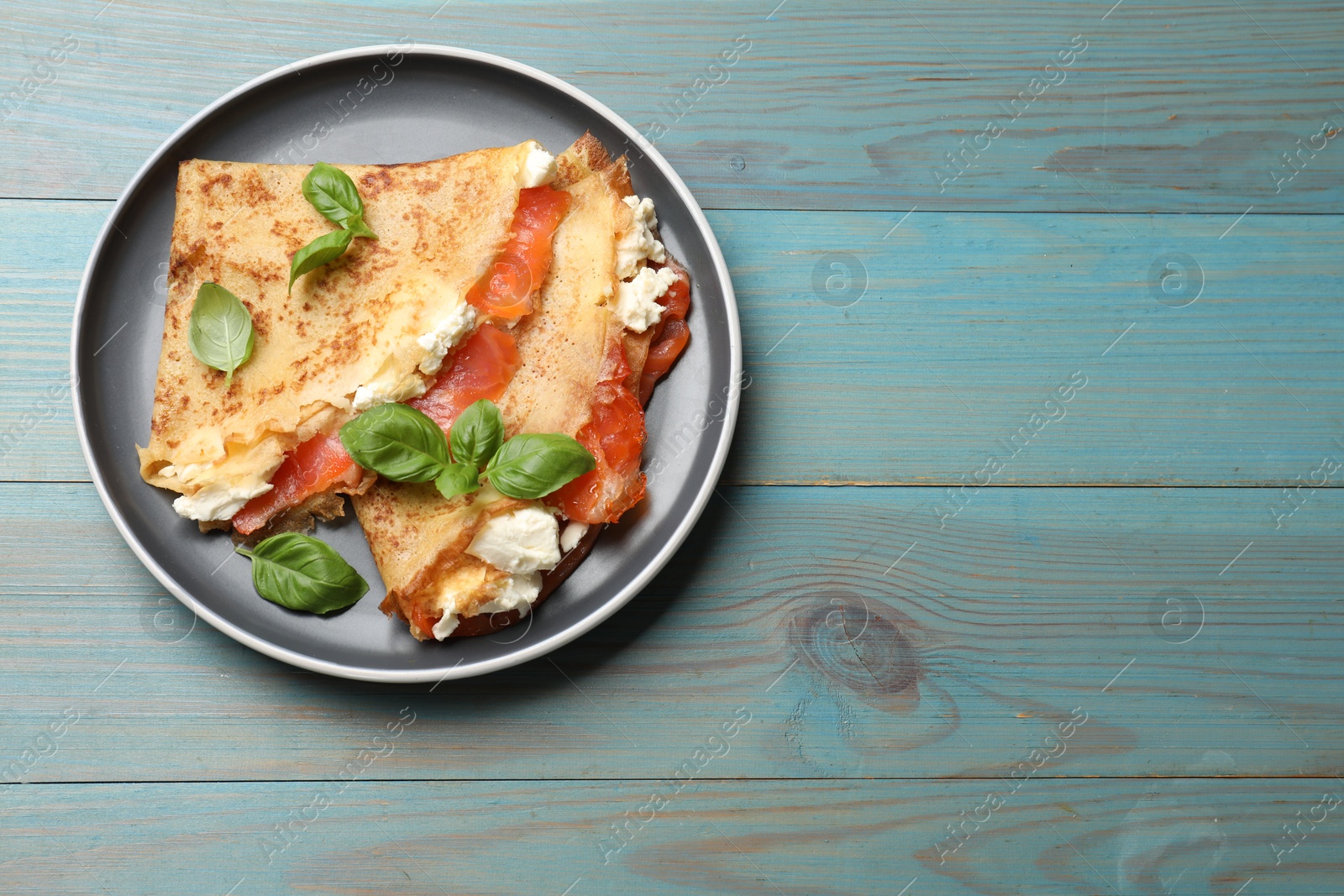 Photo of Delicious crepes with salmon, cream cheese and basil on light blue wooden table, top view. Space for text