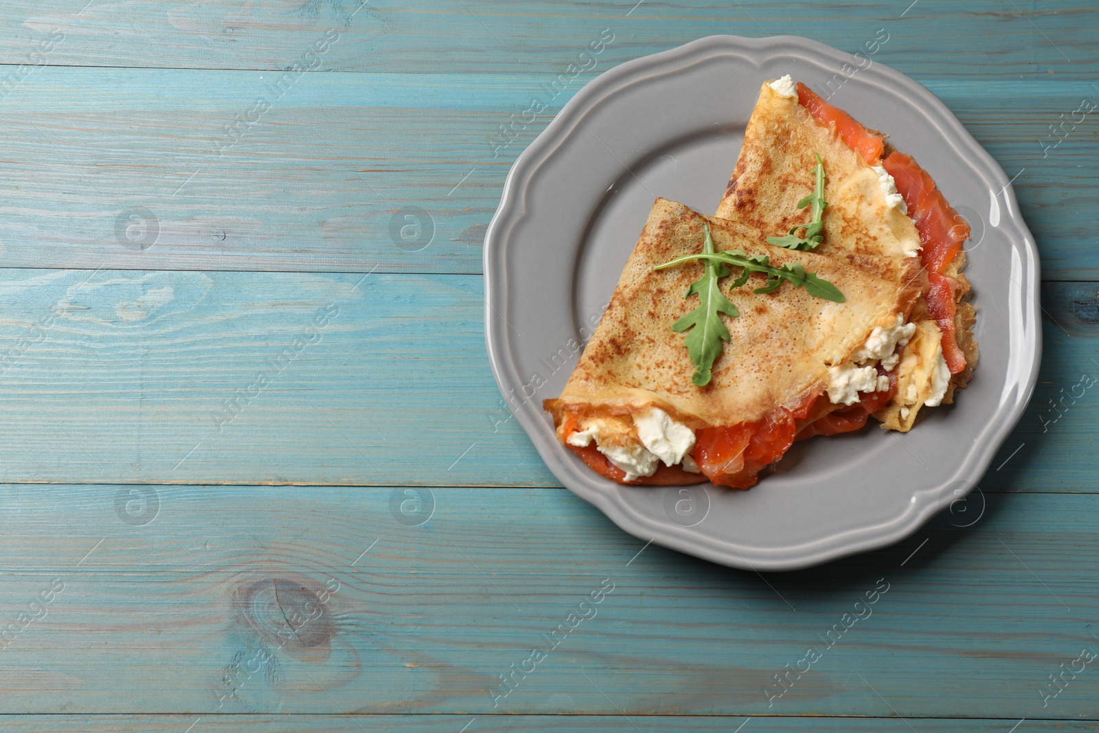 Photo of Delicious crepes with salmon, cream cheese and arugula on light blue wooden table, top view. Space for text