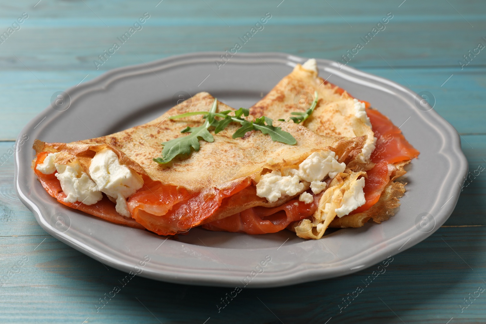 Photo of Delicious crepes with salmon, cream cheese and arugula on light blue wooden table, closeup