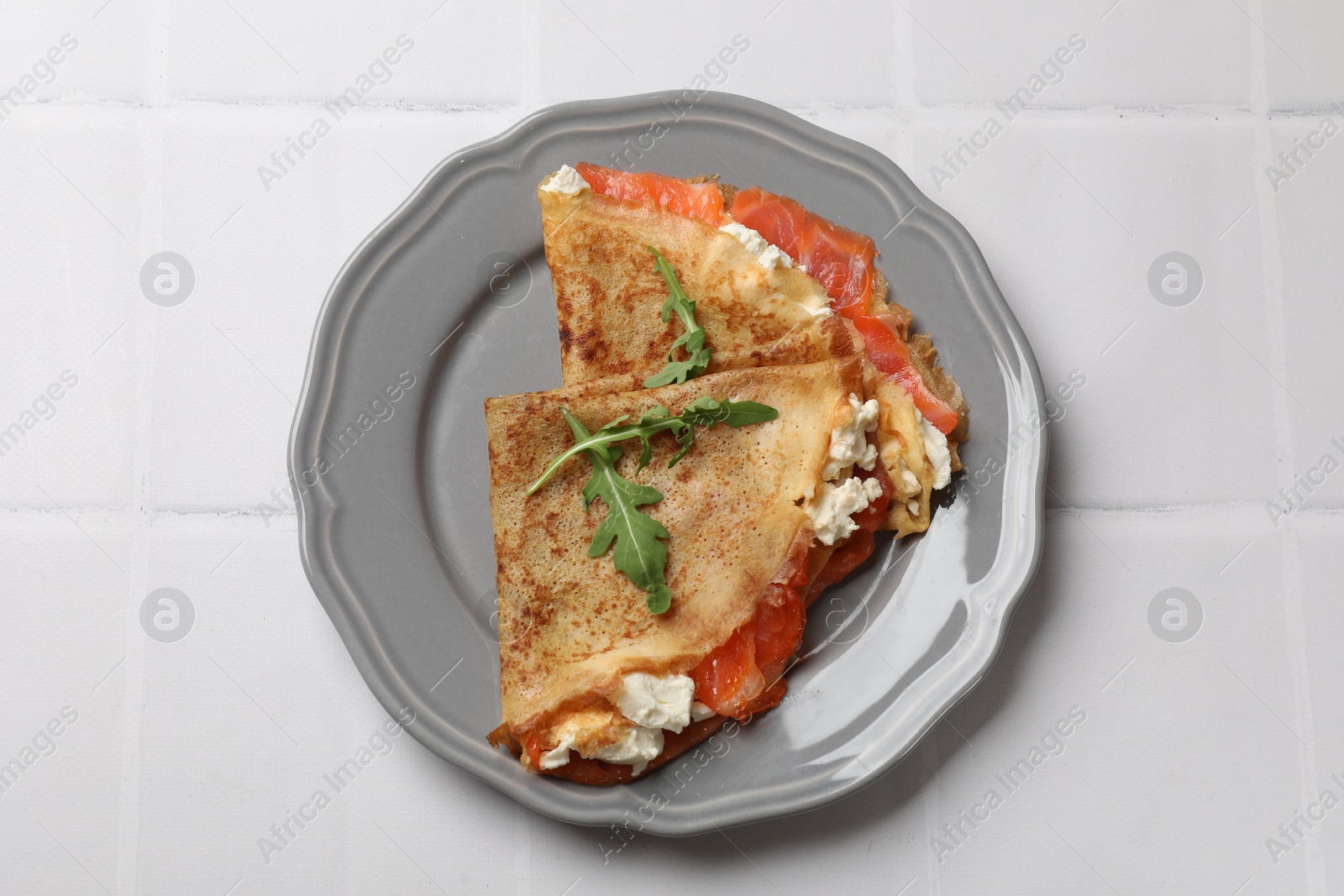 Photo of Delicious crepes with salmon, cream cheese and arugula on white tiled table, top view