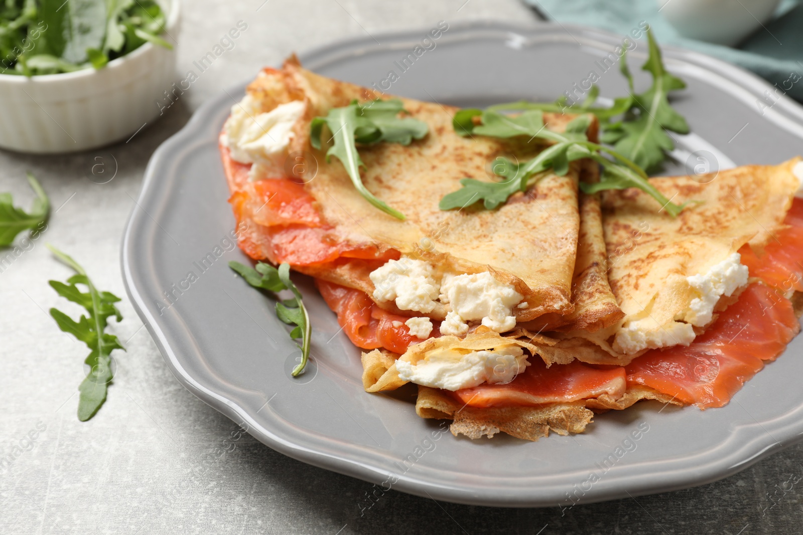 Photo of Delicious crepes with salmon, cream cheese and arugula on grey table, closeup