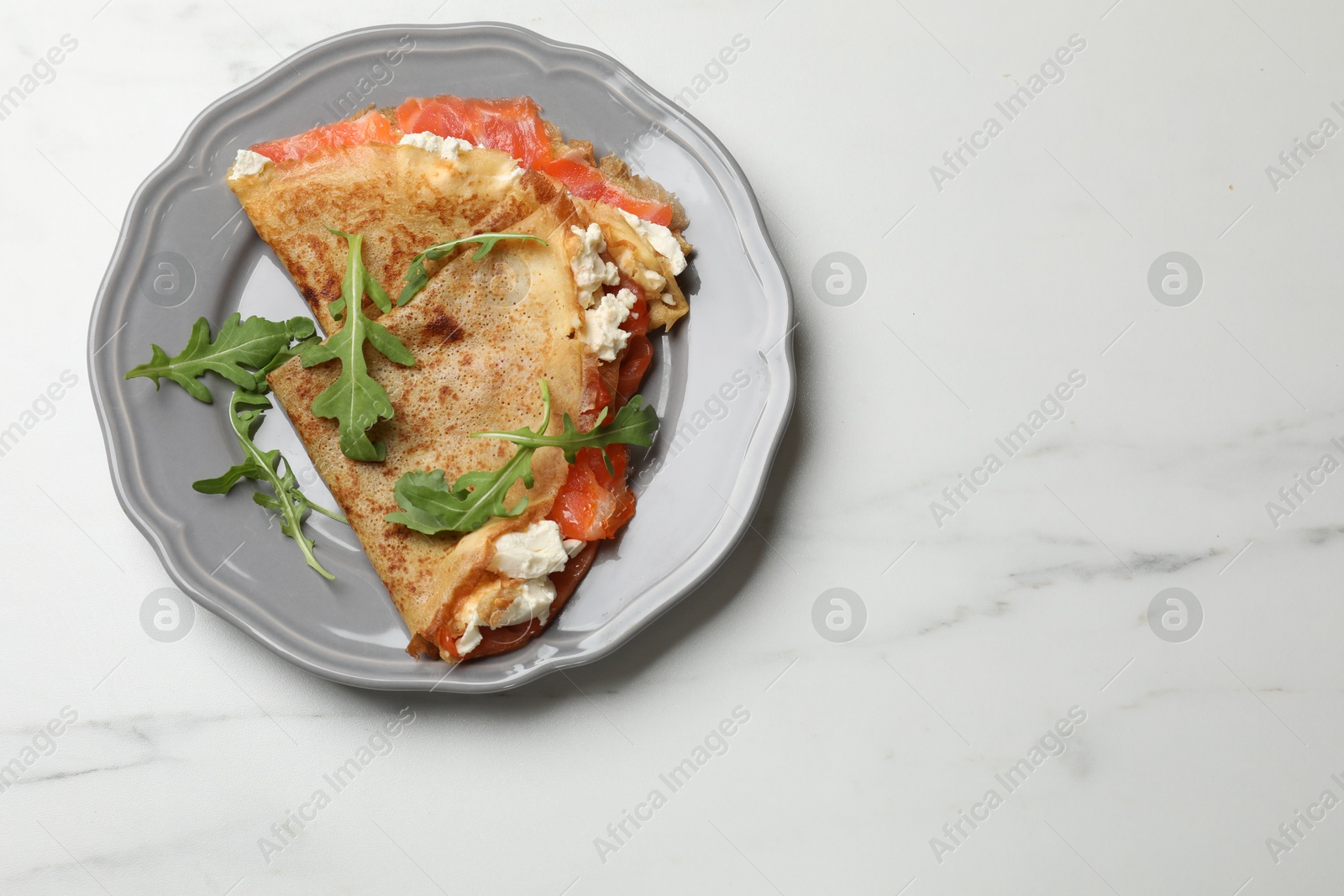 Photo of Delicious crepes with salmon, cream cheese and arugula on white marble table, top view. Space for text