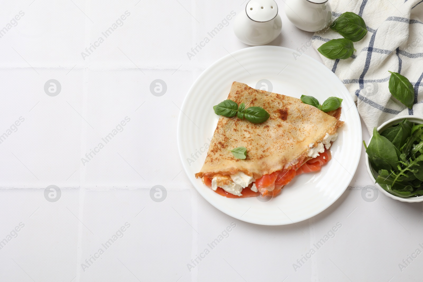 Photo of Delicious crepe with salmon, cream cheese, basil and arugula on white tiled table, flat lay. Space for text