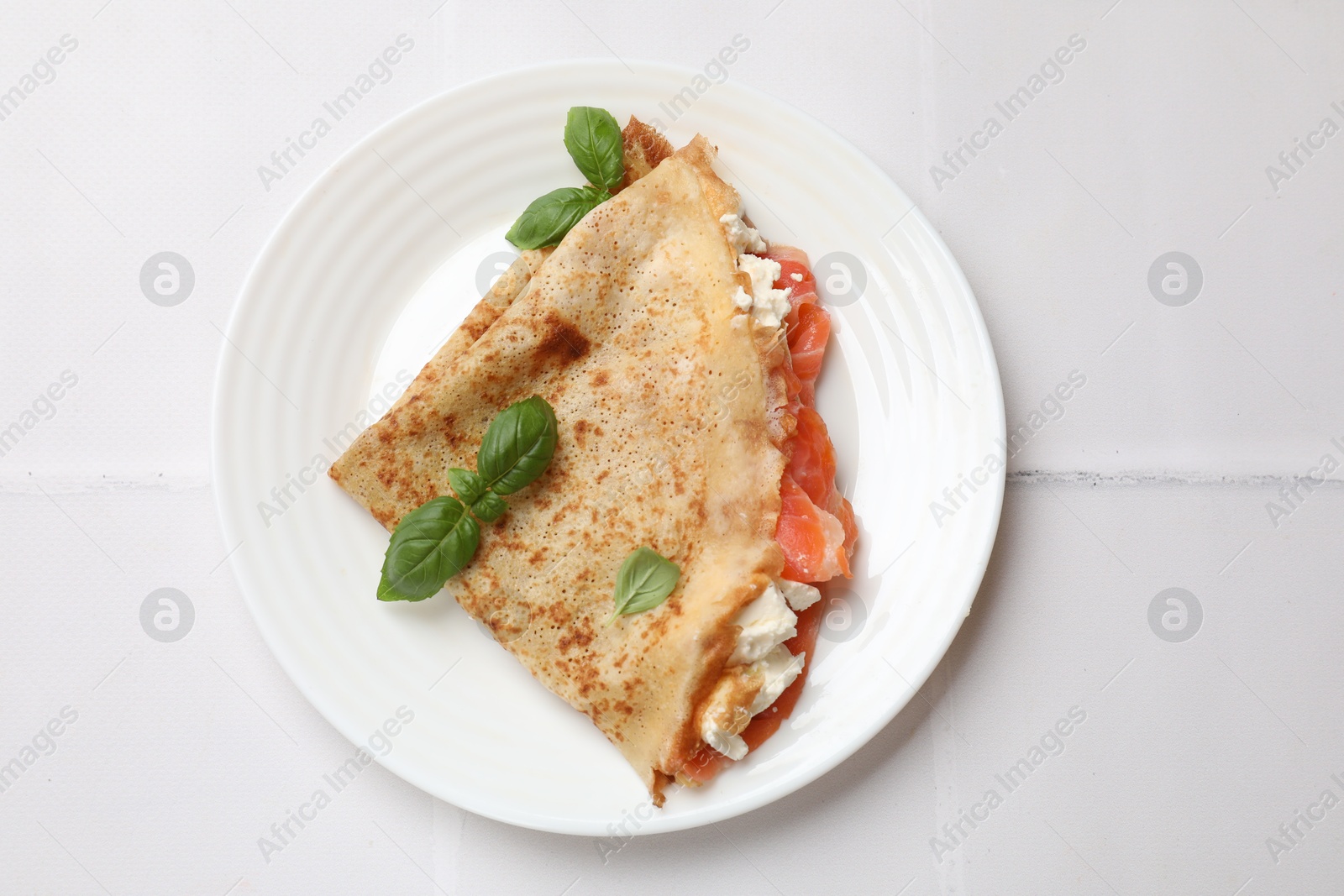 Photo of Delicious crepe with salmon, cream cheese and basil on white tiled table, top view