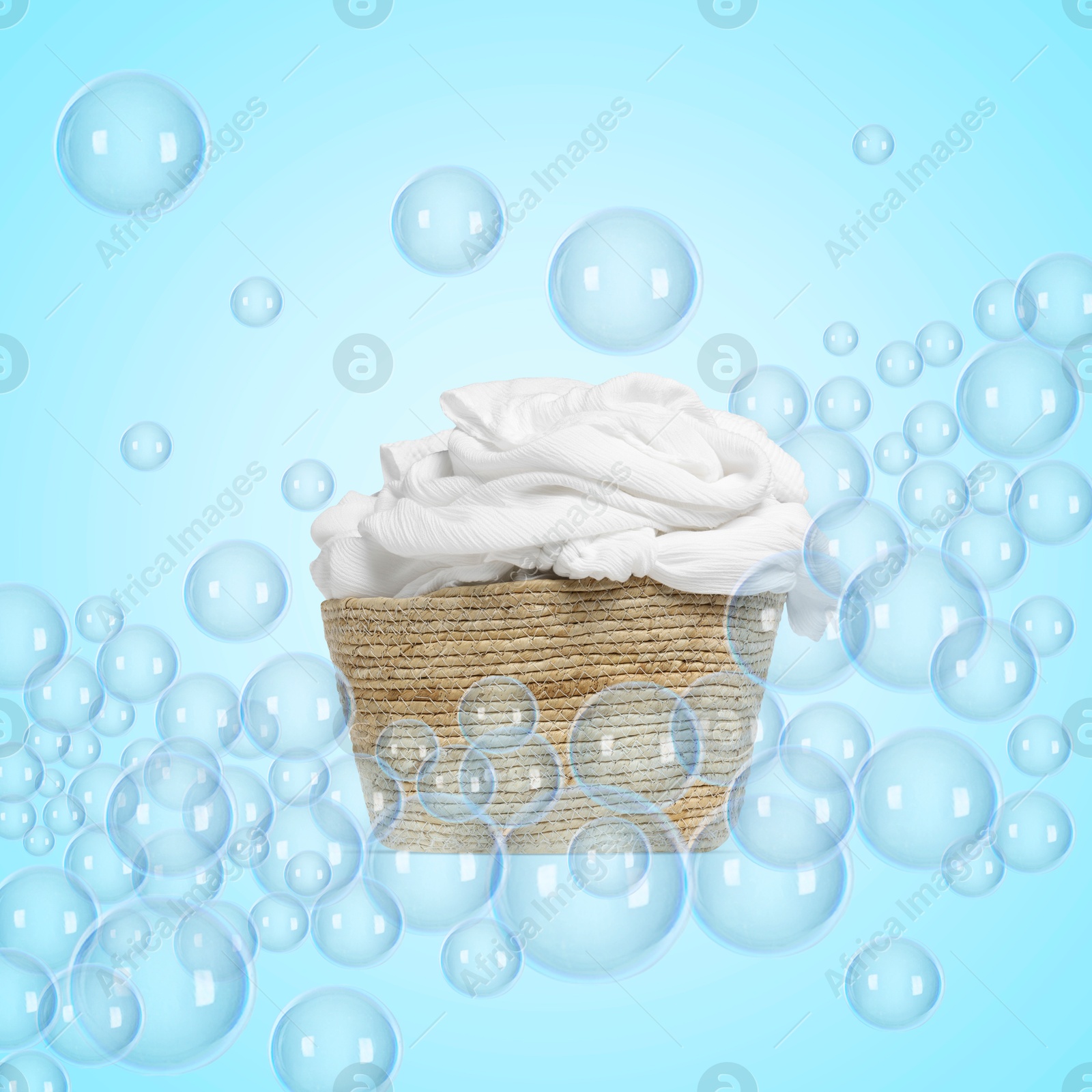 Image of Wicker basket with laundry among floating soap bubbles on light blue background