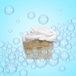 Image of Wicker basket with laundry among floating soap bubbles on light blue background