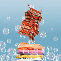 Image of Stacked laundry and flying dress among floating soap bubbles on light blue background