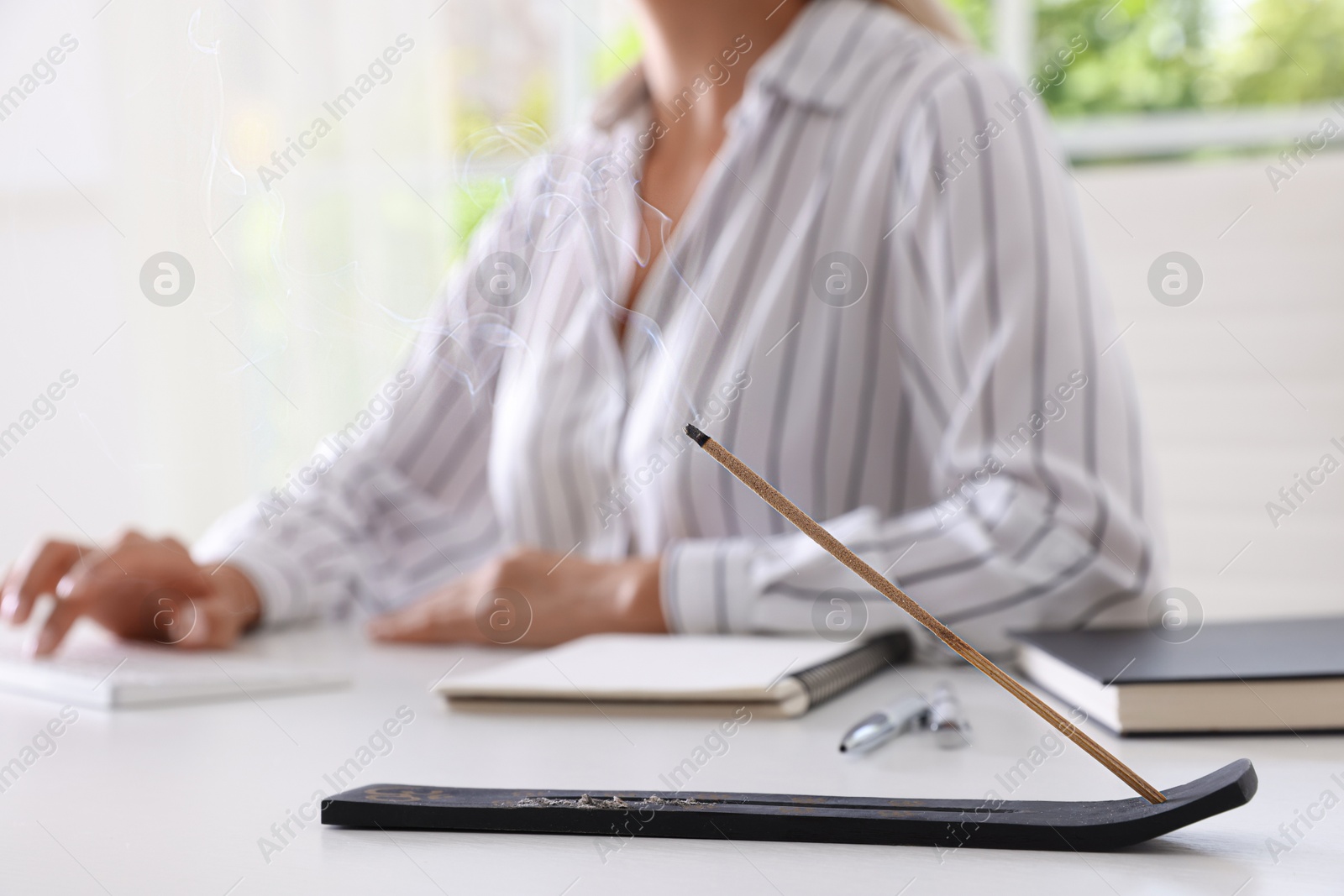 Photo of Woman working at table while incense stick smoldering in holder indoors, selective focus